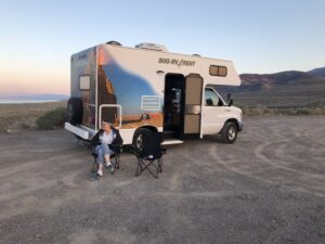 alt="person sat beside camper van at lakeside campsite"