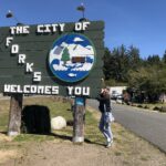 alt="person stood by the sign welcoming you to Forks"