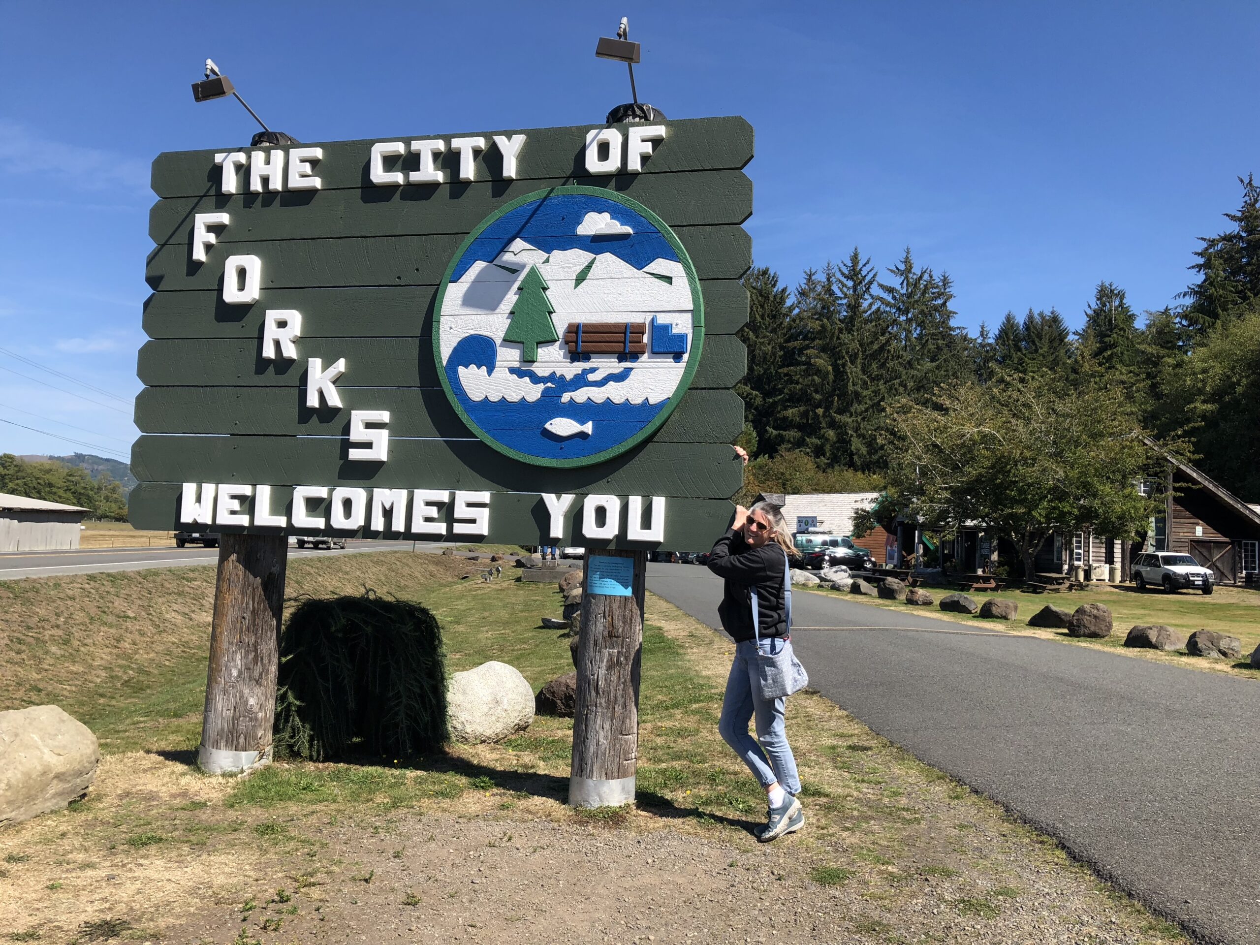 alt="person stood by the sign welcoming you to Forks"