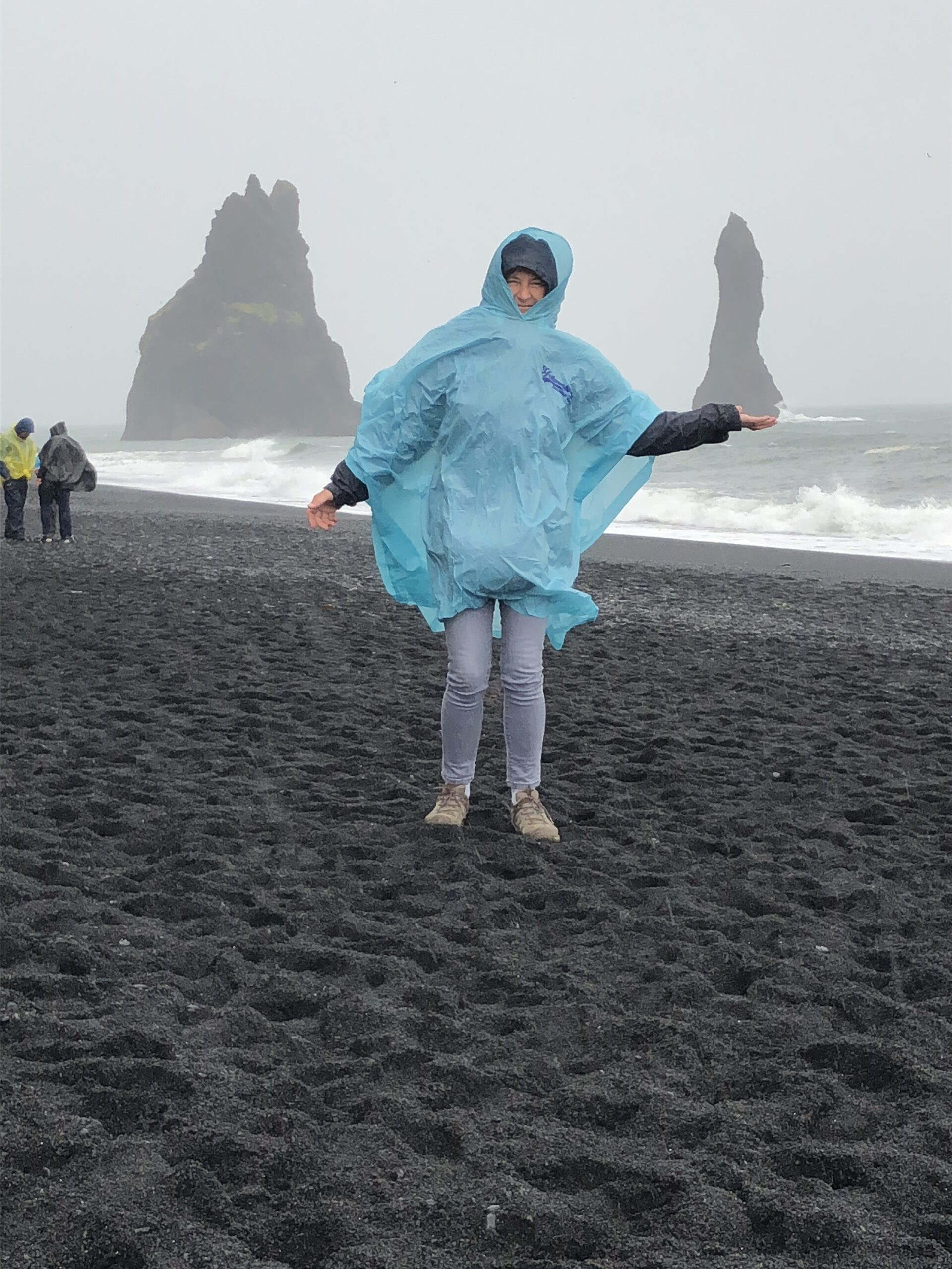 alt="person stood on the dark sand with waves & rocks in background"