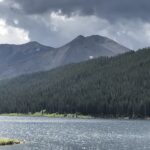alt="water with tree covered hill & mountains in background"